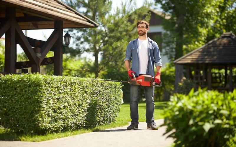 Happy bearded gardener standing carrying modern hand hedge trimmer in park low angle view of the garden